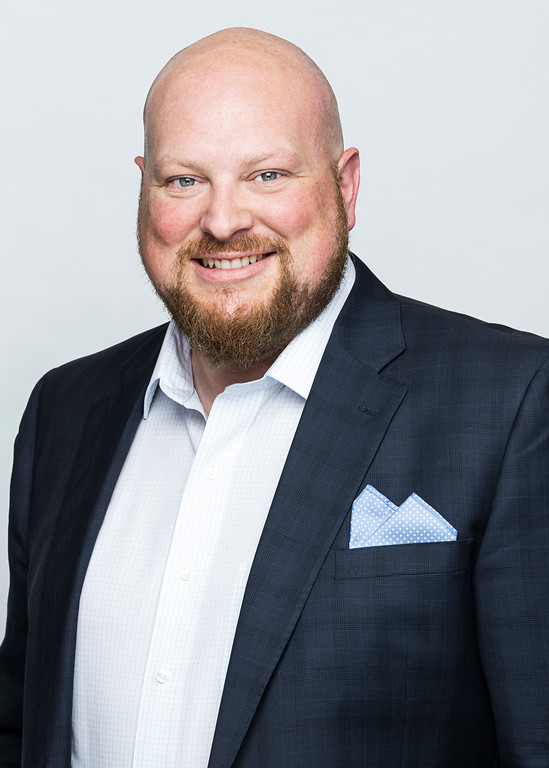 Smiling man with a bald head, beard, and light skin, wearing a dark suit jacket, white dress shirt, and a light blue pocket square.